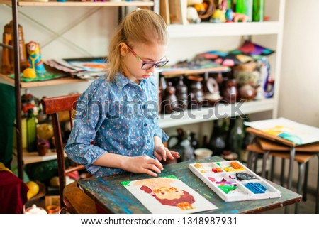 Portrait of blonde girl with big black glasses smiling happily while enjoying art and craft lesson in art school working together with other kids.