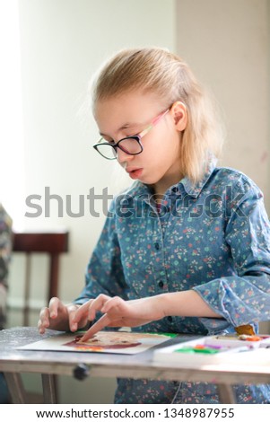Portrait of blonde girl with big black glasses smiling happily while enjoying art and craft lesson in art school working together with other kids.