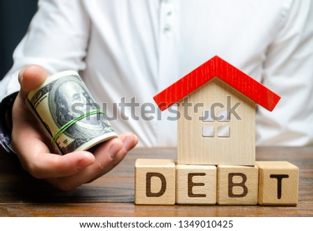 Wooden blocks with the word Debt, house and dollars in the hands of a businessman. The concept of timely payment of debt for real estate. Buying a property in debt. Mortgage. Credit for the apartment.