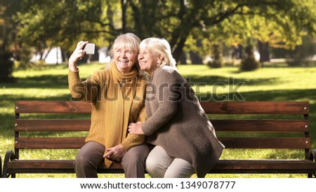 Two senior women taking selfie on cellphone, sitting on bench in park technology