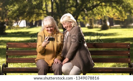 Smiling mature ladies watching photos or news on smartphone sitting in park