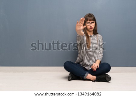 Woman with glasses sitting on the floor making stop gesture denying a situation that thinks wrong
