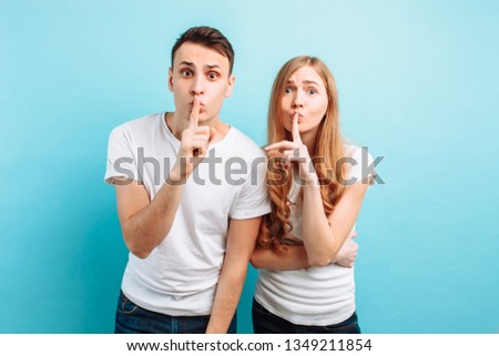 Young couple, man and woman, holding fingers on the lips of the mouth, Hush, secret, showing shhs, looking into the camera, on a blue background
