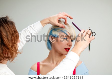 Picture showing adult woman at the hair salon. Studio shot of graceful young girl with stylish short haircut and colorful hair on gray background and hands of hairdresser.