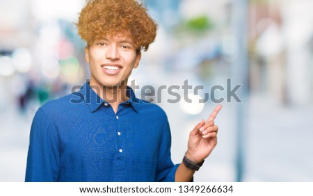 Young handsome elegant man with afro hair with a big smile on face, pointing with hand and finger to the side looking at the camera.