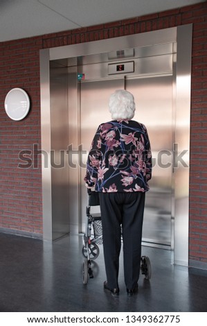 old woman with grey hair walking with her walker, in nursing home with elevator. 