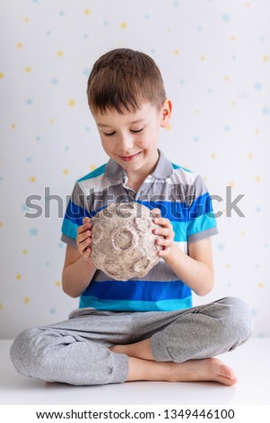 Сhild holds the moon in its hands with craters on a white starry background. A six year boy enthusiastically studies the mockup of a papier-mâché moon. Cosmonautics Day. Pre-school science education.