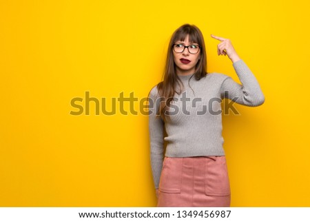 Woman with glasses over yellow wall making the gesture of madness putting finger on the head