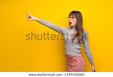 Woman with glasses over yellow wall pointing away