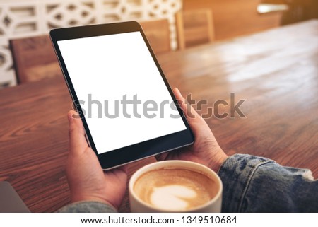 Mockup image of hands holding black tablet pc with blank white screen with coffee cup on wooden table 