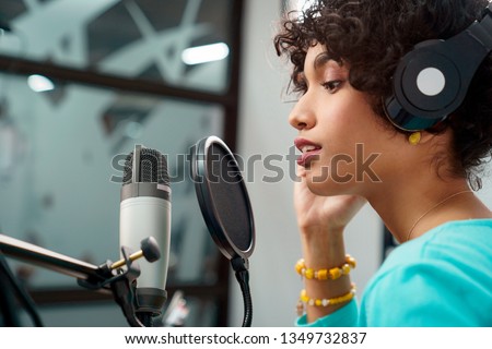 Attractive Young Black Woman Singing Into Microphone in Recording Studio Session. African American Female and podcast host with curly hair in Radio Show Talking to Microphone while wearing headphones