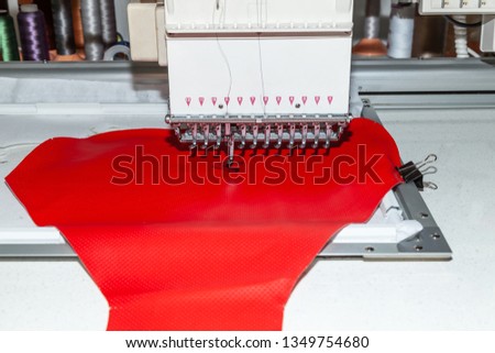 Industrial white sewing machine for embroidery makes black logo with letter S  with black thread on a red background from perforated leather
