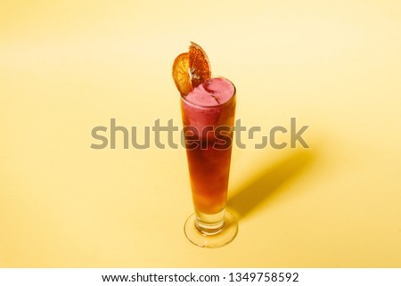 Red lemonade served in tall glass decorated with rose cream and dried orange slice on the yellow background