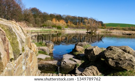 Herrington Country Park in Sunderland.  Image features trees, rock wall and a lake that is reflecting a cloudless sky on a spring/summer day.