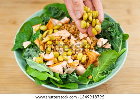 Hand adding edamame to a fresh salad bowl with lentils, shredded salmon, spinach and kale. 