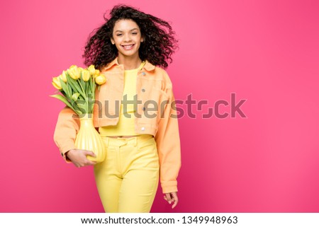smiling african american girl holding vase with yellow tulips while standing isolated on crimson