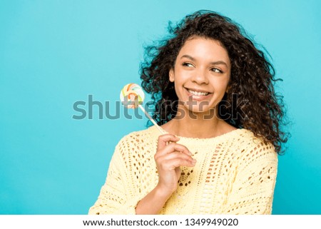 happy curly african american girl smiling while holding lollipop isolated on blue