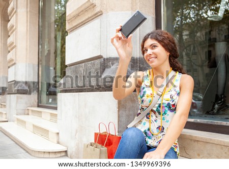 Beautiful ethnic diverse trendy tourist young woman on city holiday sitting by clothing store window using smartphone, smiling fun networking ourdoors. Technology travel leisure recreation lifestyle.