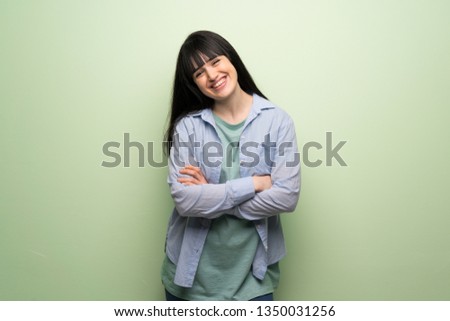 Young woman over green wall keeping the arms crossed while smiling