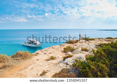 Two tiny figures of girls standing on the edge of a cliff. One white tourist ship in a bay near Cape Greco, Cyprus. Rock coastline near deep green transparent azure water. Warm day in fall