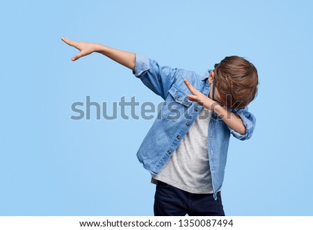 Little cool boy in glasses and denim jacket dabbing while posing on blue background  Royalty-Free Stock Photo #1350087494