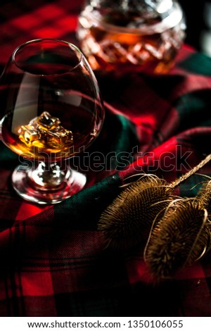 Scottish whiskey in a glass with ice on traditional red with green tartan. Crustal decanter and dry thistle. Selective focus.