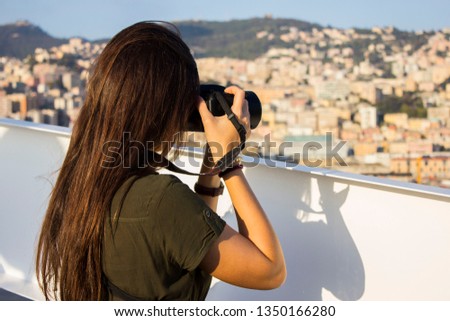 Beautiful girl with long dark hair, brunette, standing back in a green dress, holding camera, from the side of the ship is making a photograph of the view of the coastal city of Genoa in Italy.
