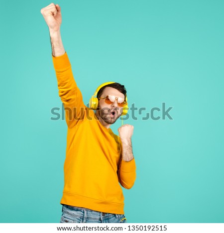 Awesome bearded young man wearing yellow sunglasses listening to music with yellow headphones feeling happy over cyan background.