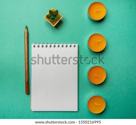 Photo flat lay layout of a blank sheet of paper in a notebook with a black pencil and a decorative cactus and orange candles on a blue-green background top view.