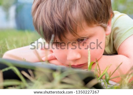Little handsome boy looking on ipad tablet and playing game at park, yard, outdoor. Kid with tablet lying on green grass. Summer holidays spending on fresh air with tablet, pc. Eye problems background
