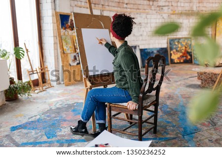 Young attractive woman with dark curly hair from back sitting on chair dreamily drawing picture on canvas spending time in big cozy art workshop with big window