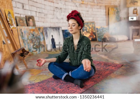 Young attractive smiling woman with closing eyes sitting on yoga mat in lotus pose and meditating spending time in cozy workshop with modern paintings on background 