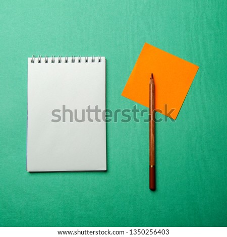Photo flat lay layout of a blank sheet of paper in a notebook with a black pencil and a colored sheet of sticker on a blue-green background, top view.