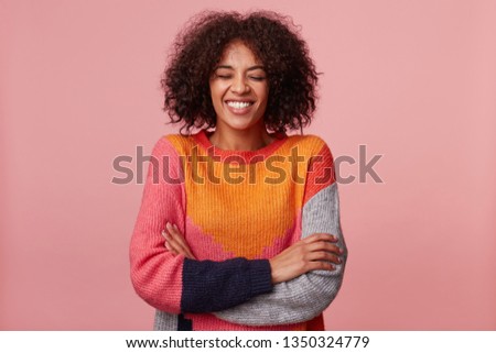 Happy joyful attractive African American with an afro hairstyle closed eyes and laughs from something funny, stand with arms crossed, wearing colorful sweater, isolated on pink Royalty-Free Stock Photo #1350324779