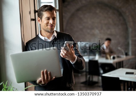 Happy businessman standing by the window in the office and reading text message on mobile phone while holding laptop. 