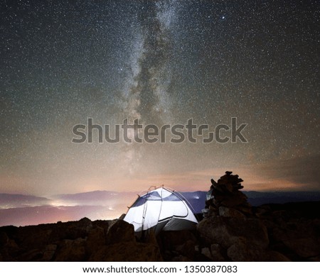Tourist camping at night on the top of rocky mountain. Illuminated tent under incredibly beautiful night sky full of stars and Milky way. On background amazing starry sky, mountains and luminous city
