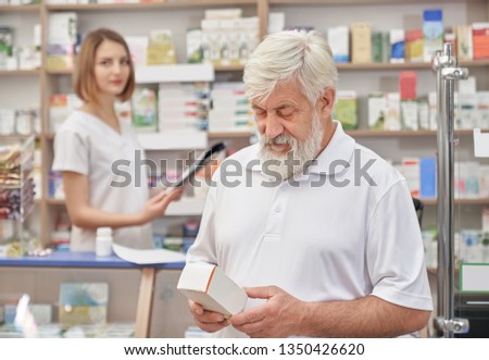 Elderly man choosing medicaments in drugstore, thinking. Bearded pensioner in white t shirt holding medical box, looking down. Female pharmacist standing at counter in pharmacy.