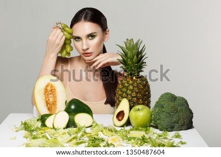 Picture of beautiful young brunette woman with fruits and vegetables on the table, holding green grapes in hand isolated on grey background