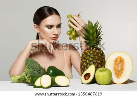 Picture of beautiful young brunette woman with fruits and vegetables on the table, holding green grapes in hand isolated on grey background