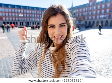 Beautiful young caucasian woman happy and excited in Plaza Mayor Madrid holding the camera and taking a photo of herself. Looking cheerful and joyful. In tourism, European city, travel in Europe.