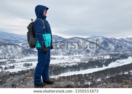 A young girl in bright winter clothes with a backpack is standing on top of a mountain smiling and looking at other mountains on a cloudy winter day. hiking in the mountains.