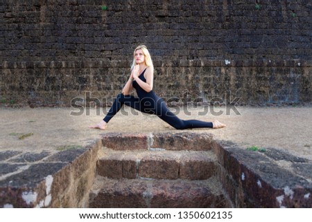 Blonde girl doing yoga in an ancient Portuguese Fort in India. The black stones were overgrown with grass. Healthy lifestyle with yoga