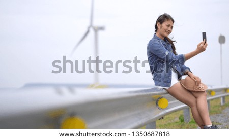 Asian women are photographing selfie from a mobile phone, with the background being a wind turbine and a meadow.

