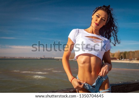 Young beautiful athletic woman in a short vest and blue ripped jeans posing against a beach on a warm summer day near the sea. Summer concept. Holiday travel