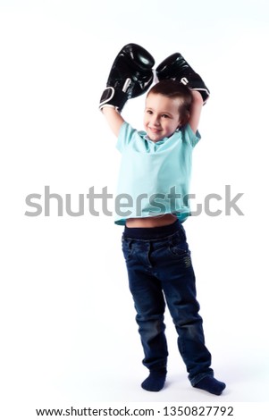 A little boy with dark hair in blue jeans, a blue polo shirt in black and white boxing gloves is having fun, showing biceps and considering himself a winner on a white isolated background in a  studio