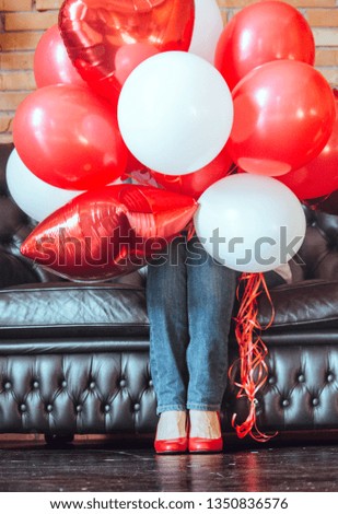 Unknown girl hidden in red and white helium balloons on black sofa. Colorful balloons and women legs in red shoes. Birthday decoration. Happy birthday concept. Red and white balloons. 
