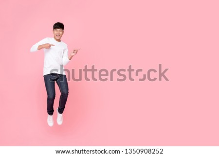 Young cheerful energetic handsome Asian man jumping with hands pointing to copy space aside studio shot isolated on pink background
