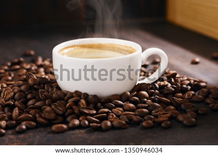 Cup of hot coffee with steam and roasted coffee beans. Dark background, warm sunlight. Royalty-Free Stock Photo #1350946034