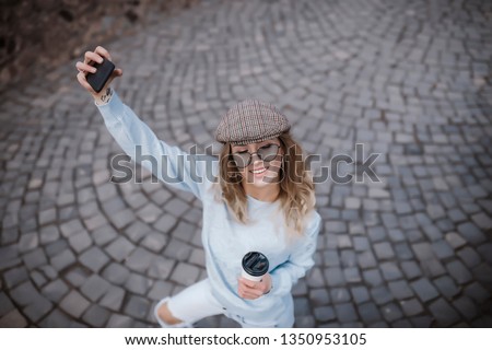 Portrait of wonderful white female model with bright makeup expressing energy in good day in Europe. Lovely curly woman in stylish hat making selfie while walking past old building. 
