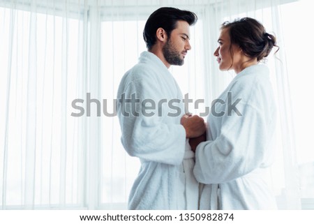 handsome bearded man holding hands with happy woman in white bathrobe in hotel 